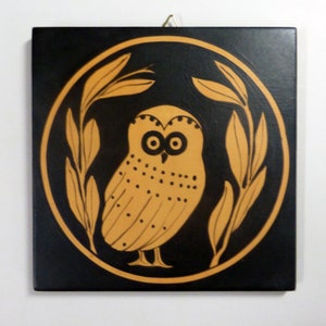 May include: Square black wall art featuring a stylized owl in a circle, surrounded by olive branches. The owl and branches are in a light brown color, contrasting against the black background. A gold-colored wire is attached for hanging.