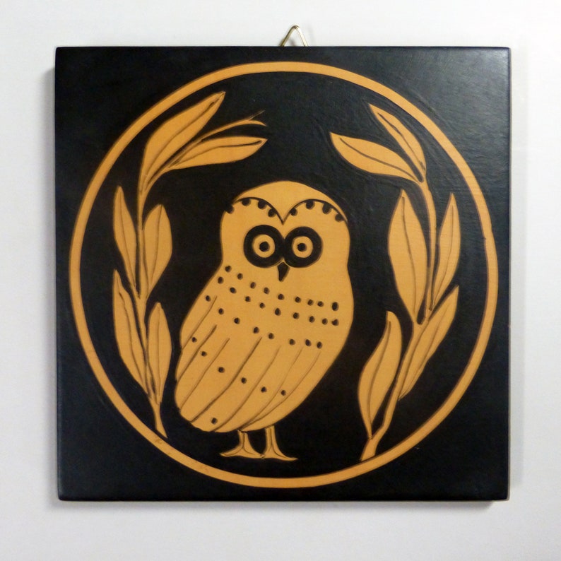 May include: Square black wall art featuring a stylized owl in a circle, surrounded by olive branches. The owl and branches are in a light brown color, contrasting against the black background. A gold-colored wire is attached for hanging.
