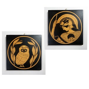 May include: Two square, black wall plaques with gold-colored illustrations. One features an owl within a laurel wreath, the other depicts a figure in a helmet and armor. Both are framed by a gold circle.