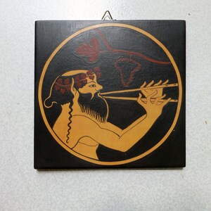 May include: Square, black wooden plaque with a circular design featuring a figure playing a wind instrument. The figure is in tan with red accents, set against a black background with a gold border. A red vine design is above the figure.