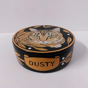 May include: A round, black trinket box with a detailed illustration of a tabby cat on the lid. The cat has green eyes and is surrounded by gold-colored leaves. The name "DUSTY" is printed in gold on the side of the box.