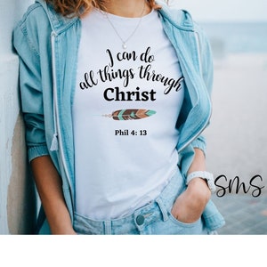 Puede incluir: Camiseta blanca con el texto "Puedo hacerlo todo por medio de Cristo" y un diseño de pluma marrón y turquesa. El texto "Phil 4:13" está debajo del diseño.
