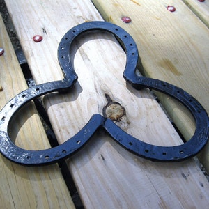 May include: A black metal horseshoe trivet, made from three horseshoes welded together in a clover shape. The trivet is sitting on a wooden surface.