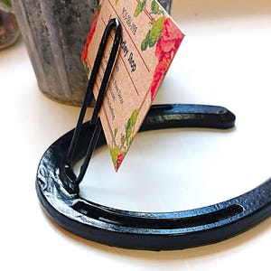 May include: Black horseshoe-shaped business card holder with a clip. The card holder is holding a business card with the text "Valley Shop" and a floral design.