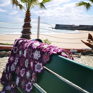 May include: A purple and white crocheted blanket with a flower pattern draped over a green bench. The bench is in front of a beach with a palm tree and a stone wall in the background.