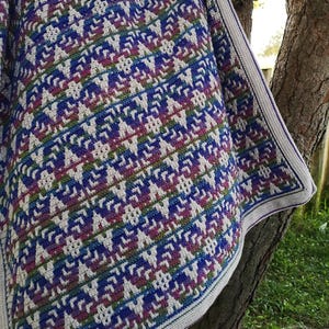 Puede incluir: Manta de ganchillo hecha a mano con un patrón detallado en tonos azules, morados, verdes y crema. La manta presenta un diseño geométrico repetitivo y un borde color crema. La manta está sobre una rama de árbol.