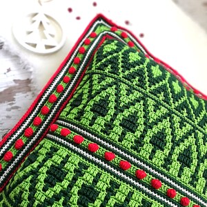 May include: Green and red crocheted pillow with a geometric pattern. The pillow has a white border with red pom-poms.