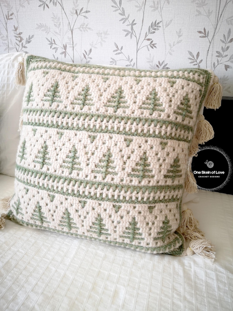 May include: A crocheted cushion with a green and white geometric pattern featuring stylised trees. The cushion has fringe tassels on all four corners. The cushion is on a white bedspread with a floral patterned wallpaper in the background.