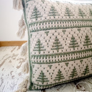 May include: A green and white crocheted cushion with a fringe border and a pattern of stylised pine trees. The cushion is on a whitewashed wooden surface. The text "One Skein of Love Crochet Designs" is visible in the bottom right corner of the image.