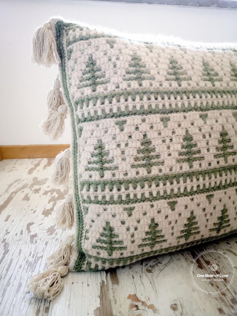 May include: A green and white crocheted cushion with a fringe border and a pattern of stylised pine trees. The cushion is on a whitewashed wooden surface. The text "One Skein of Love Crochet Designs" is visible in the bottom right corner of the image.