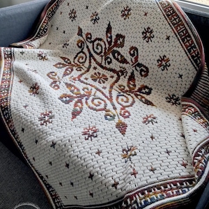May include: A cream-colored, crocheted blanket with a detailed floral and geometric pattern in shades of brown, red, and gold. The blanket has a textured appearance and a decorative border. The blanket is draped over a dark gray couch.