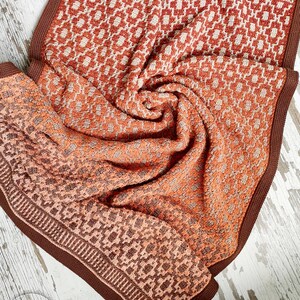 Puede incluir: Una manta de crochet marr&oacute;n y naranja con un patr&oacute;n geom&eacute;trico. La manta est&aacute; doblada y tiene un borde marr&oacute;n.