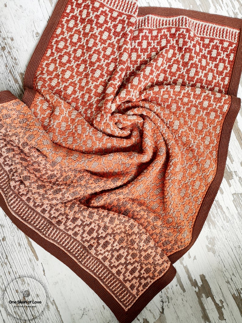 Puede incluir: Una manta de crochet marr&oacute;n y naranja con un patr&oacute;n geom&eacute;trico. La manta est&aacute; doblada y tiene un borde marr&oacute;n.