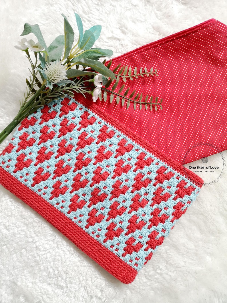 Puede incluir: Una bolsa de cremallera de ganchillo con un patr&oacute;n geom&eacute;trico rojo y azul. La bolsa est&aacute; hecha con un solo ovillo de hilo y tiene un forro de lunares rojos. La bolsa est&aacute; sobre una superficie blanca y peluda.