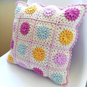 Puede incluir: Una almohada de crochet con fondo blanco y diseños de flores de colores en rosa, amarillo y azul. La almohada tiene un borde de crochet rosa.