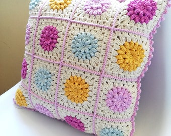 May include: A crocheted pillow with a white background and colorful flower designs in pink, yellow, and blue. The pillow has a pink crocheted border.