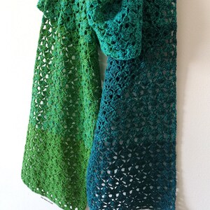 May include: A green and teal crochet scarf with a gradient pattern. The scarf is made with a delicate open stitch and has a fringed edge.