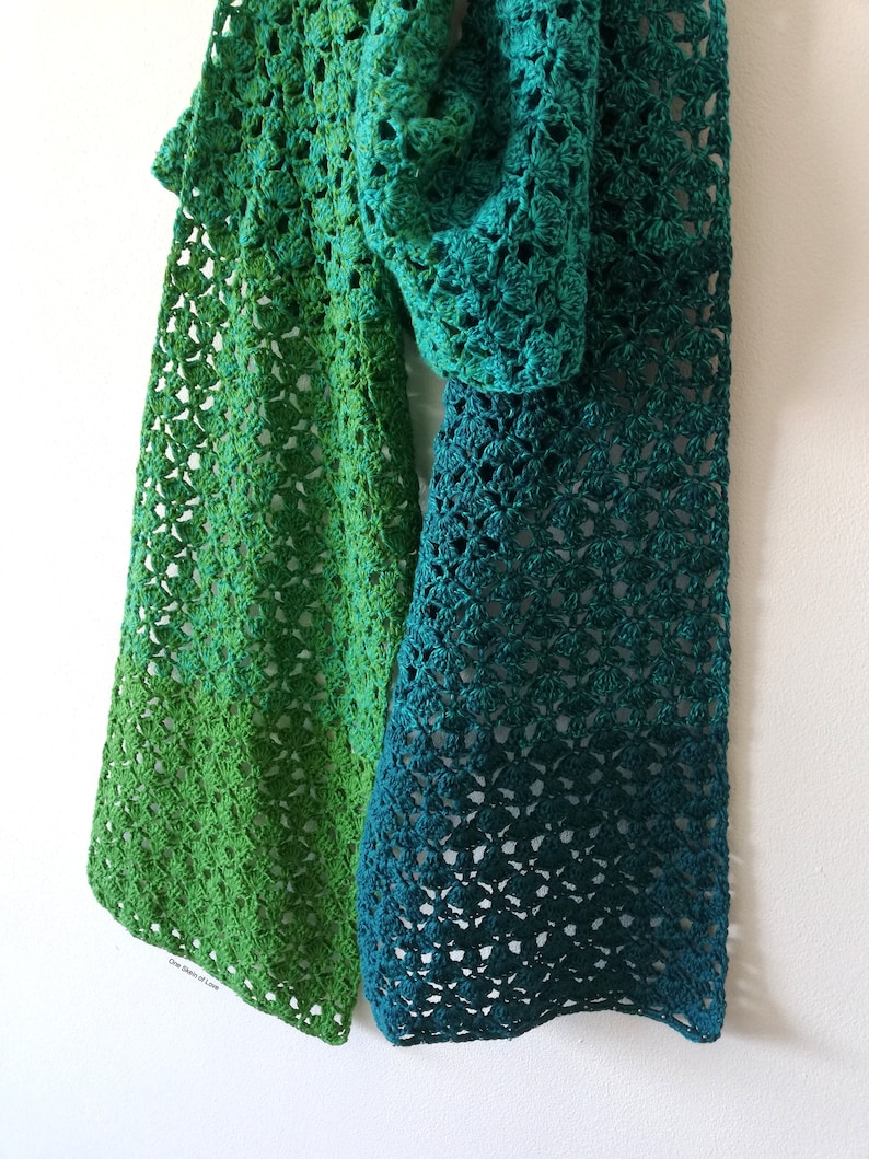 May include: A green and teal crochet scarf with a gradient pattern. The scarf is made with a delicate open stitch and has a fringed edge.