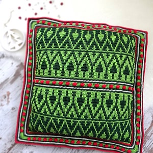 May include: Green and red crocheted pillow with a geometric pattern. The pillow has a white border and red pom-poms around the edge. The text "One Stitch of Love" is visible in the bottom left corner.