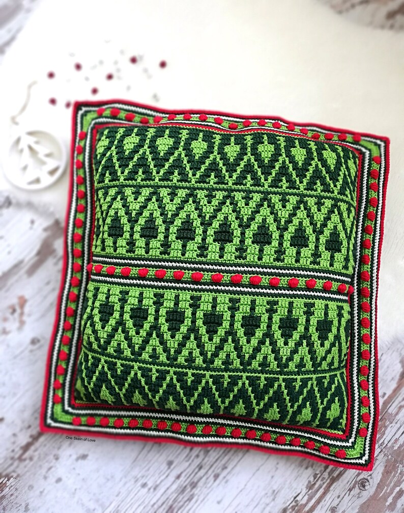 May include: Green and red crocheted pillow with a geometric pattern. The pillow has a white border and red pom-poms around the edge. The text "One Stitch of Love" is visible in the bottom left corner.