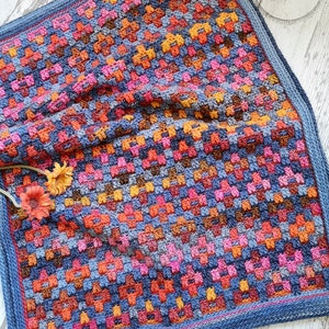 Może przedstawiać: Kolorowy koc szydełkowy z niebieskim obramowaniem. Koc wykonany jest z różnych kolorów, w tym pomarańczowego, różowego, czerwonego i brązowego. Koc jest złożony na pół i leży na drewnianej powierzchni. Tekst "One Skein of Love Crochet Designs" jest widoczny w prawym górnym rogu obrazu.