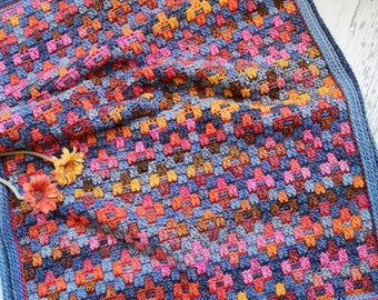 May include: A colourful crochet blanket with a blue border. The blanket is made with a variety of colours, including orange, pink, red, and brown. The blanket is folded in half and is lying on a wooden surface. The text "One Skein of Love Crochet Designs" is visible in the top right corner of the image.
