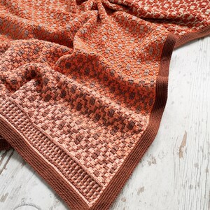 Puede incluir: Una manta de ganchillo con un patr&oacute;n de cuadros marrones y naranjas. La manta tiene un borde marr&oacute;n y est&aacute; doblada sobre una superficie de madera blanca. El texto "One Skein of Love Crochet Designs" es visible en la esquina inferior izquierda de la imagen.