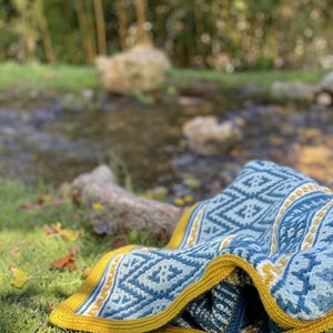 May include: A blue and yellow crocheted blanket with a geometric pattern is lying on green grass near a stream. The blanket has a white border and a yellow fringe. The text "One Skein of Love" is visible in the bottom right corner of the image.