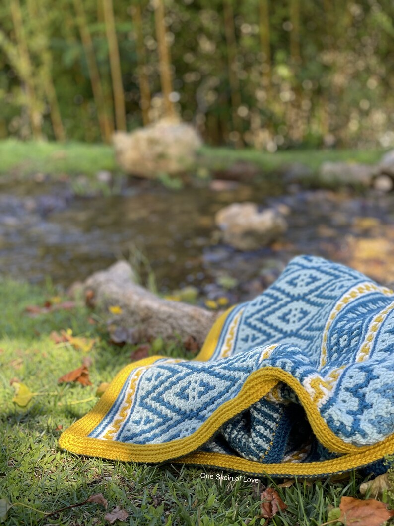 May include: A blue and yellow crocheted blanket with a geometric pattern is lying on green grass near a stream. The blanket has a white border and a yellow fringe. The text "One Skein of Love" is visible in the bottom right corner of the image.