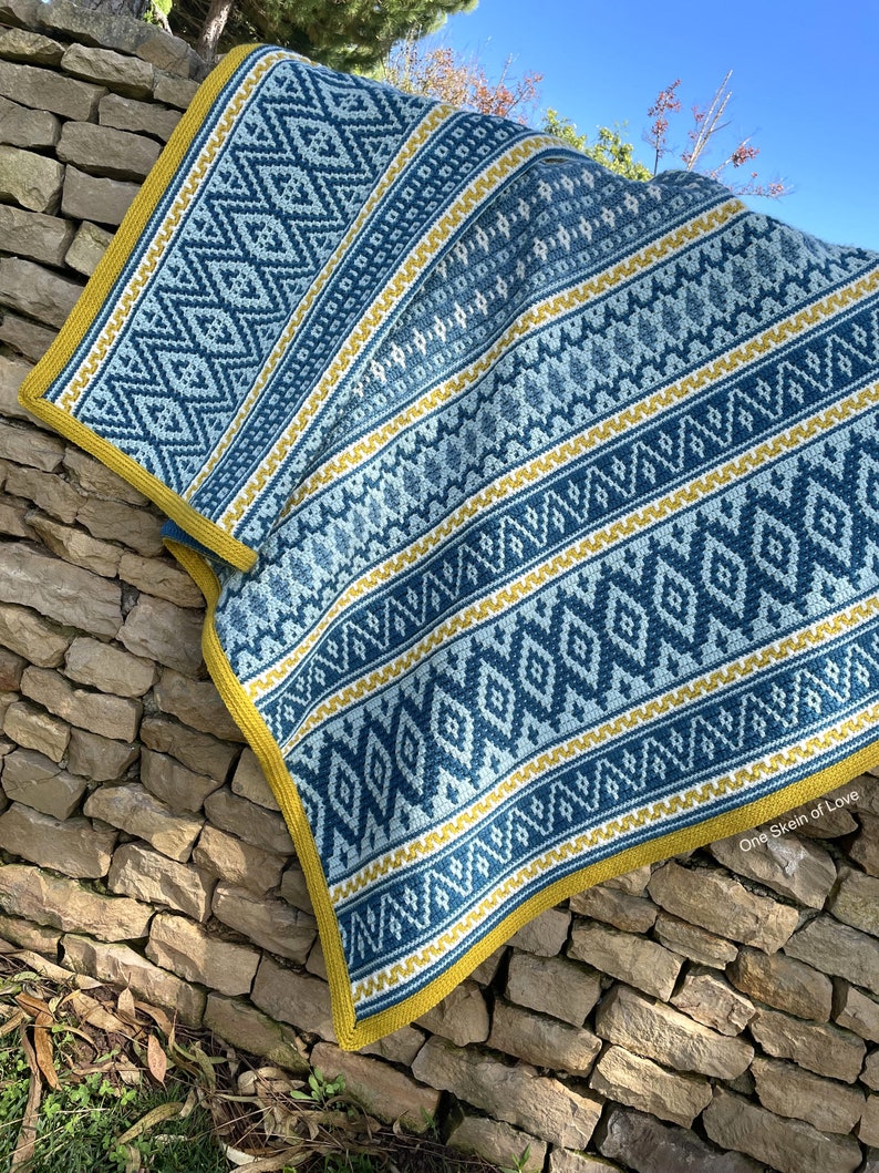 May include: A blue and yellow crocheted blanket with a geometric pattern. The blanket is folded and draped over a stone wall. The text "One Skein of Love" is visible in the bottom right corner.