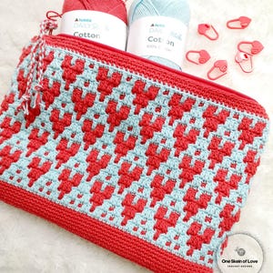 Puede incluir: Una bolsa de cremallera de crochet roja y azul con un patr&oacute;n de coraz&oacute;n. La bolsa est&aacute; hecha con hilo "Hobbia Daily Stitch Cotton". La bolsa est&aacute; sobre una superficie blanca y peluda con marcadores de punto en forma de coraz&oacute;n rojo.