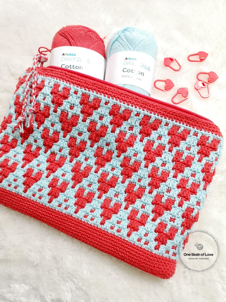 Puede incluir: Una bolsa de cremallera de crochet roja y azul con un patr&oacute;n de coraz&oacute;n. La bolsa est&aacute; hecha con hilo "Hobbia Daily Stitch Cotton". La bolsa est&aacute; sobre una superficie blanca y peluda con marcadores de punto en forma de coraz&oacute;n rojo.