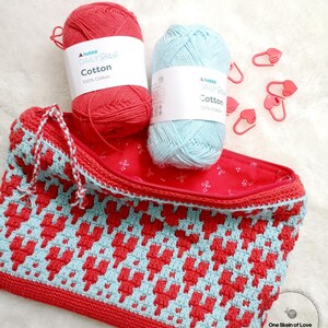 Puede incluir: Una bolsa de cremallera de crochet roja y azul con un patr&oacute;n de coraz&oacute;n. La bolsa est&aacute; hecha con dos ovillos de hilo, uno rojo y uno azul, y tiene una cremallera. La bolsa est&aacute; sentada sobre una superficie blanca y peluda con marcadores de punto en forma de coraz&oacute;n rojo.