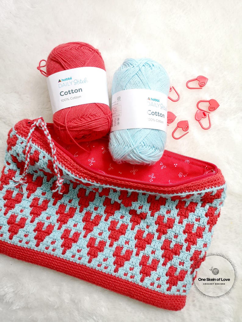 Puede incluir: Una bolsa de cremallera de crochet roja y azul con un patr&oacute;n de coraz&oacute;n. La bolsa est&aacute; hecha con dos ovillos de hilo, uno rojo y uno azul, y tiene una cremallera. La bolsa est&aacute; sentada sobre una superficie blanca y peluda con marcadores de punto en forma de coraz&oacute;n rojo.