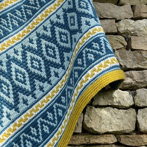 May include: A crocheted throw blanket with a geometric pattern in shades of blue, white, and yellow. The blanket is draped over a stone wall.
