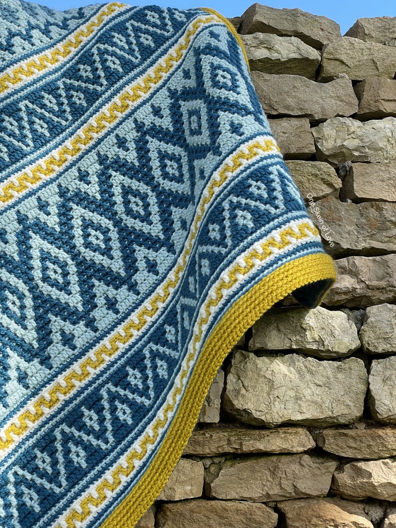 May include: A crocheted throw blanket with a geometric pattern in shades of blue, white, and yellow. The blanket is draped over a stone wall.