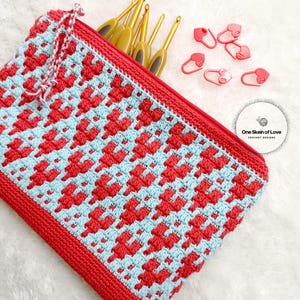 Puede incluir: Una bolsa de cremallera de crochet roja y azul con un patr&oacute;n de coraz&oacute;n. La bolsa est&aacute; llena de agujas de crochet y marcadores de punto de coraz&oacute;n rojo. El texto "One Skein of Love Crochet Designs" es visible en la esquina inferior derecha de la imagen.