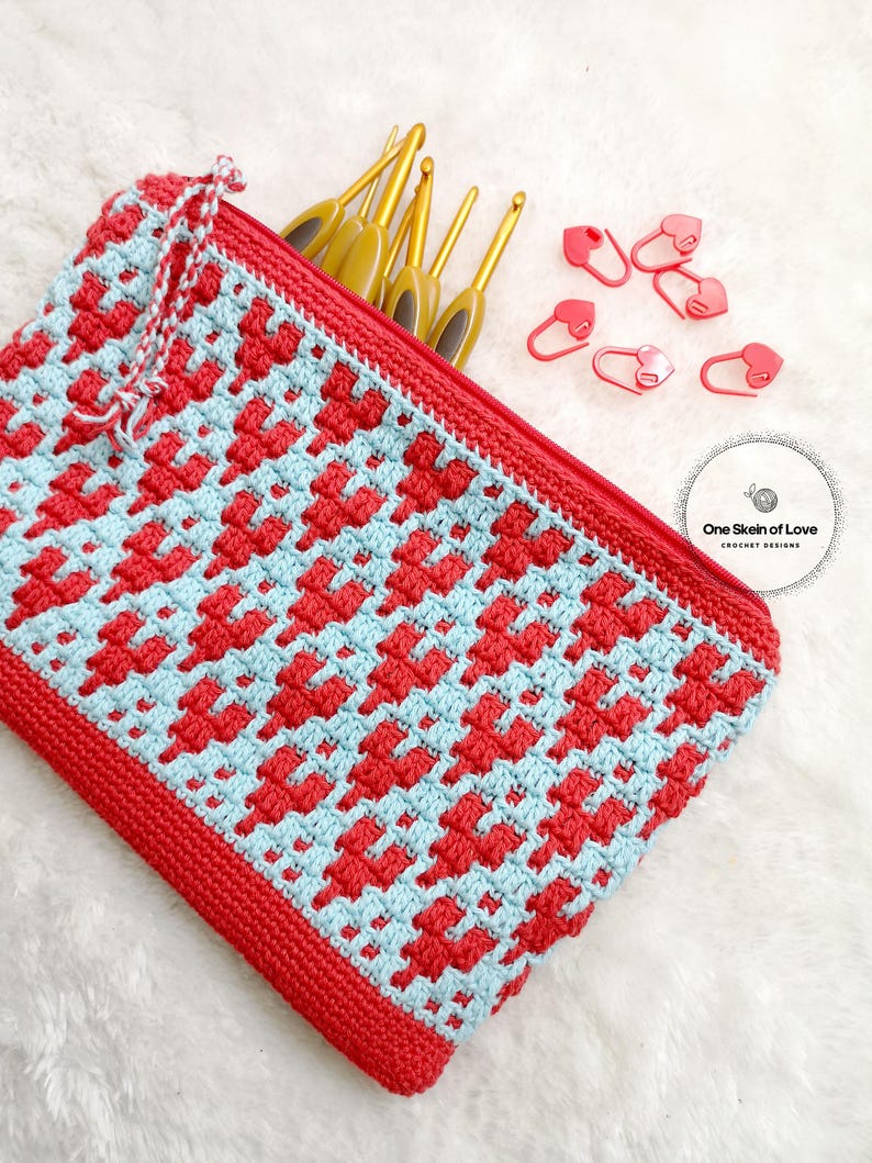 Puede incluir: Una bolsa de cremallera de crochet roja y azul con un patr&oacute;n de coraz&oacute;n. La bolsa est&aacute; llena de agujas de crochet y marcadores de punto de coraz&oacute;n rojo. El texto "One Skein of Love Crochet Designs" es visible en la esquina inferior derecha de la imagen.