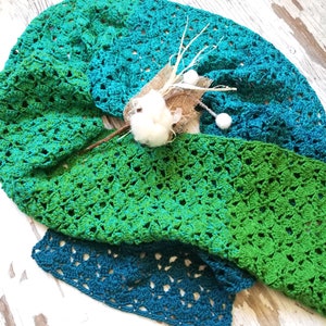 May include: A green and teal crochet shawl with a delicate open stitch pattern. The shawl is folded and draped over a rustic wooden surface.