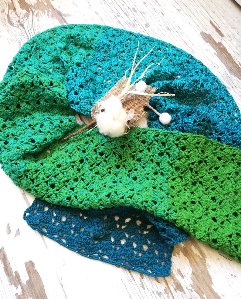 May include: A green and teal crochet shawl with a delicate open stitch pattern. The shawl is folded and draped over a rustic wooden surface.