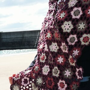 May include: A crocheted shawl featuring a floral pattern in shades of pink, brown, and purple. The shawl is crafted with a delicate stitch and has a flowing, feminine design.