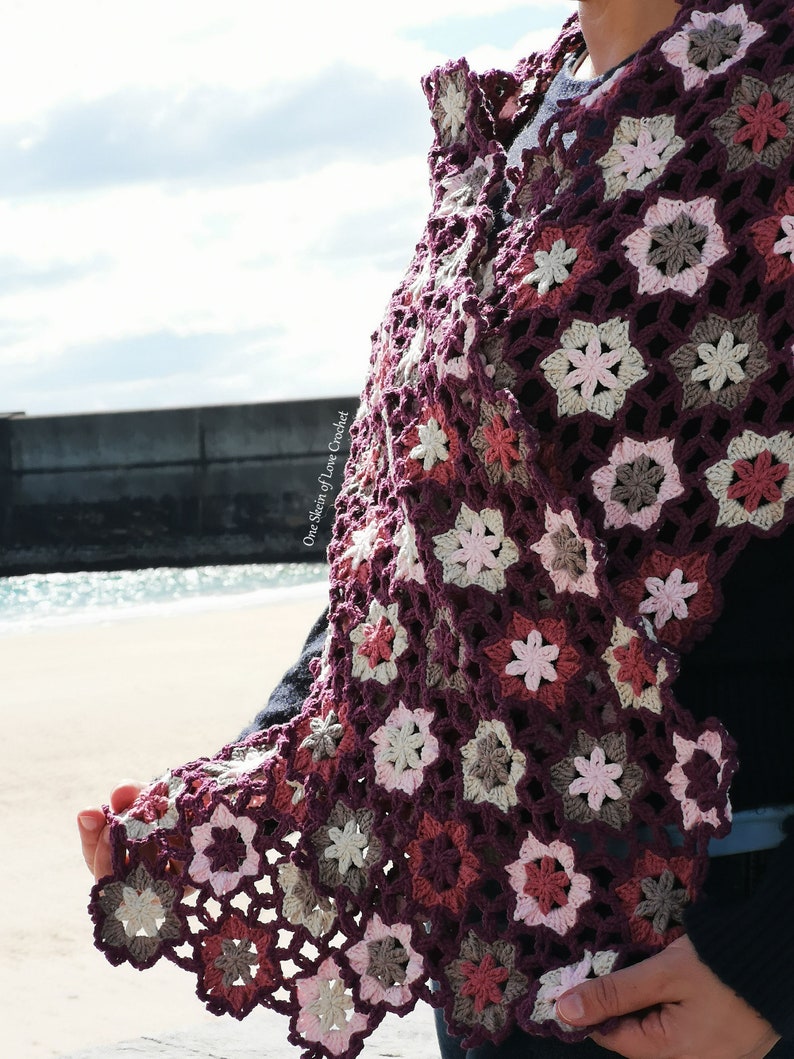 May include: A crocheted shawl featuring a floral pattern in shades of pink, brown, and purple. The shawl is crafted with a delicate stitch and has a flowing, feminine design.