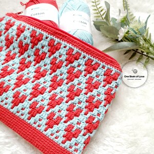 Puede incluir: Una bolsa de cremallera de ganchillo con un patr&oacute;n geom&eacute;trico en rojo y azul. La bolsa est&aacute; hecha con hilo de algod&oacute;n y tiene un cierre de cremallera. La bolsa est&aacute; sobre una superficie blanca y esponjosa con dos ovillos de lana en el fondo.