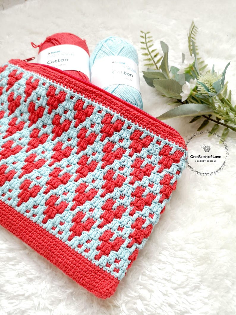 Puede incluir: Una bolsa de cremallera de ganchillo con un patr&oacute;n geom&eacute;trico en rojo y azul. La bolsa est&aacute; hecha con hilo de algod&oacute;n y tiene un cierre de cremallera. La bolsa est&aacute; sobre una superficie blanca y esponjosa con dos ovillos de lana en el fondo.