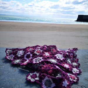 May include: A purple, pink and grey crocheted blanket with a starburst pattern. The blanket is lying on a concrete surface with a beach and ocean in the background. The text "One Stitch of Love Crochet" is visible on the blanket.