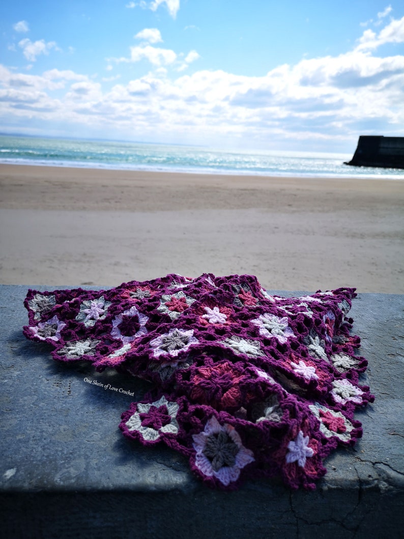 May include: A purple, pink and grey crocheted blanket with a starburst pattern. The blanket is lying on a concrete surface with a beach and ocean in the background. The text "One Stitch of Love Crochet" is visible on the blanket.