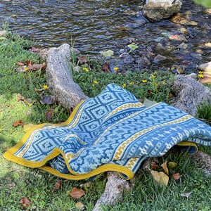 May include: A blue and yellow crocheted blanket with a geometric pattern is draped over a tree branch near a stream. The blanket has a yellow border.