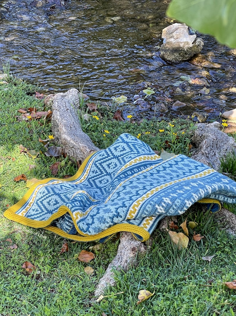 May include: A blue and yellow crocheted blanket with a geometric pattern is draped over a tree branch near a stream. The blanket has a yellow border.