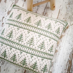 May include: A white and green crocheted cushion with a fringed border. The cushion features a repeating pattern of green stylised Christmas trees.