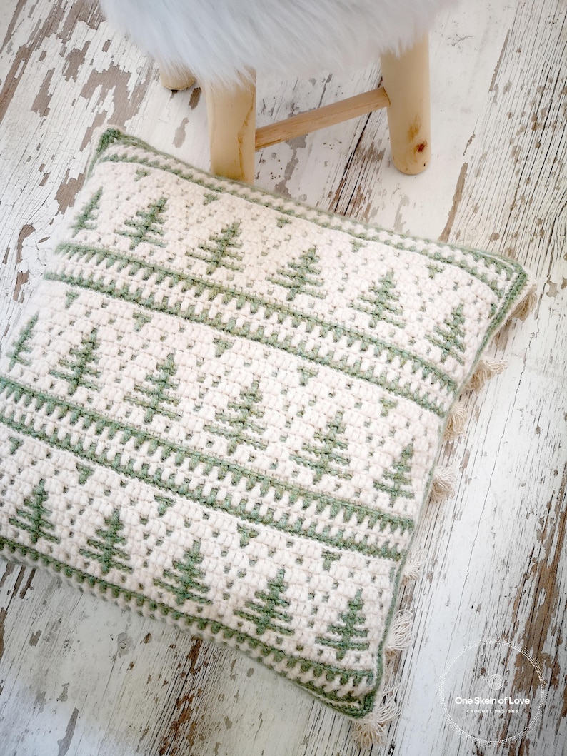 May include: A white and green crocheted cushion with a fringed border. The cushion features a repeating pattern of green stylised Christmas trees.